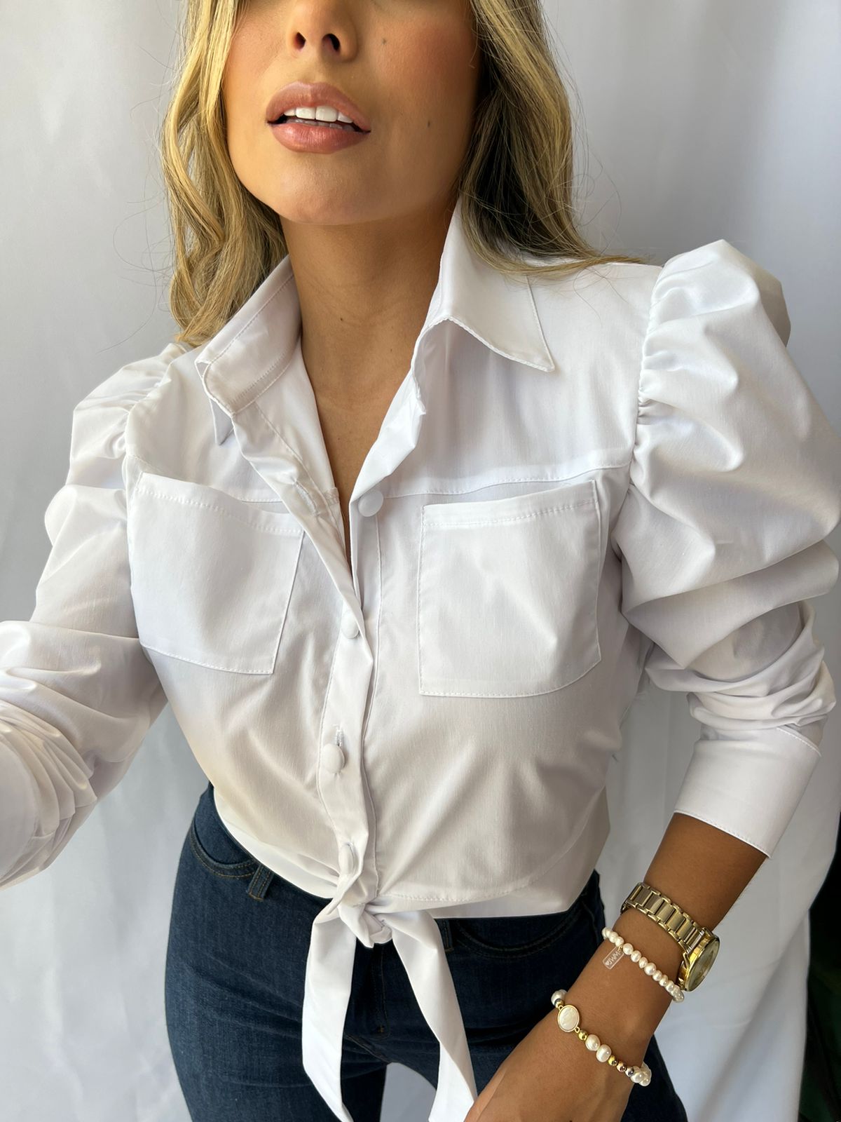 Mujer con blusa blanca de manga larga, con bolsillos y un nudo frontal, ideal para un look elegante y moderno. Disponibilidad en KIKE RODRIGUEZ.