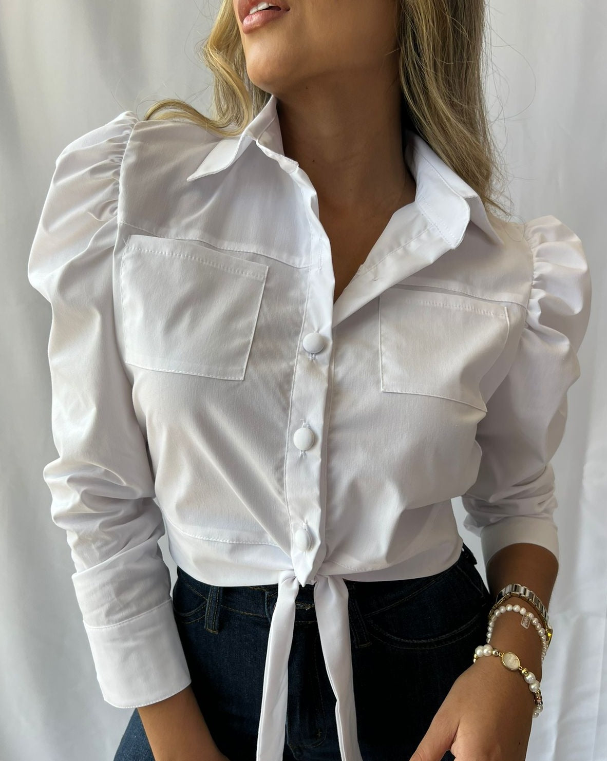 Mujer con blusa blanca de manga larga, con bolsillos y un nudo frontal, ideal para un look elegante y moderno. Disponibilidad en KIKE RODRIGUEZ.