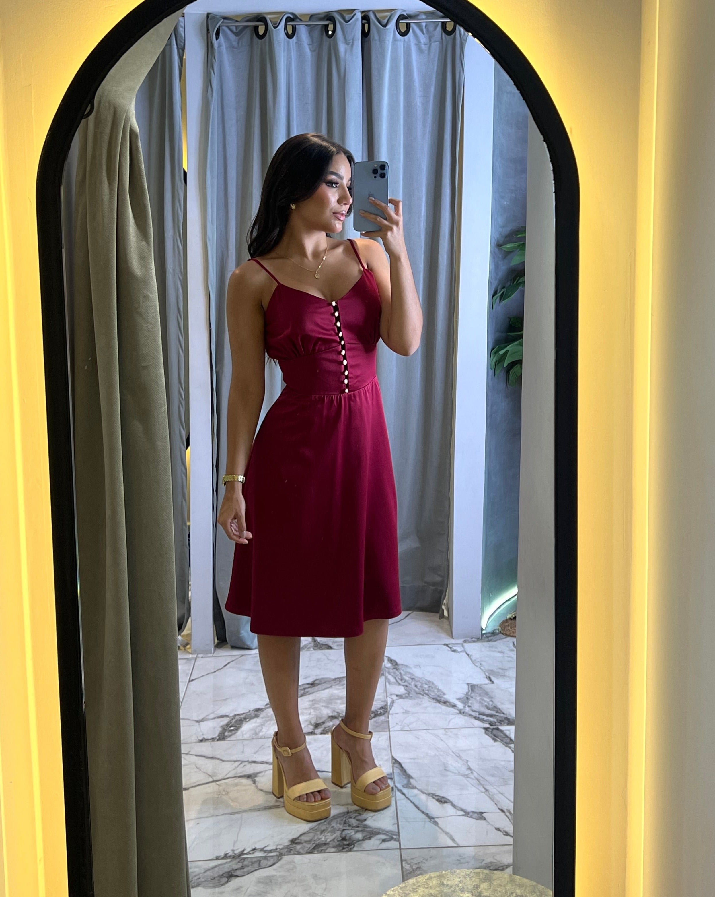 Mujer posando frente al espejo con un vestido corto en color rojo vino, tirantes finos y botones frontales, perfecto para eventos casuales y formales.